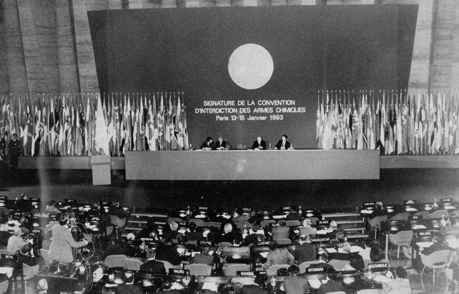 The historic black and white photography shows four people at a high table and several others attending the event. Letters on the wall say: Signature de la convention d'interdiction des armes chimiques, Paris 13-15 Janvier 1993.